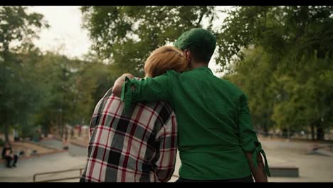 A-blonde-girl-with-a-short-haircut-in-a-plaid-shirt-leaned-on-the-shoulder-of-her-girlfriend-with-a-green-short-haircut-in-a-Green-shirt-sitting-on-the-fence-in-a-skate-park-in-the-summer.-Happy-LGBT-couple-during-their-date