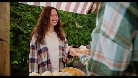 Over-the-shoulder,-a-happy-guy-in-a-green-checkered-shirt-buys-pastries-from-a-brunette-girl-in-a-checkered-shirt-in-a-shop-during-a-fair-near-the-city-against-the-backdrop-of-green-coniferous-trees-in-summer