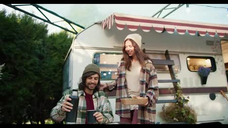 A-happy-brunette-girl-in-a-white-hat-and-a-tick-on-her-shirt-takes-out-baked-goods-for-her-boyfriend-who-pours-tea-from-a-thermos-and-sits-at-a-table-near-a-trailer-in-a-camp-outside-the-city-during-a-picnic-in-the-summer