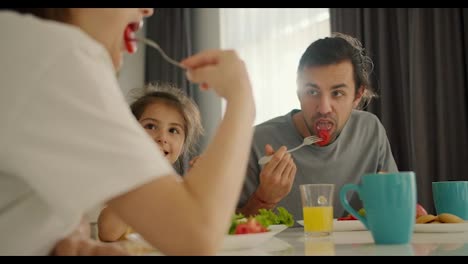 Close-up-shot:-happy-brunette-man-eats-salad-during-his-breakfast-together-with-his-little-daughter-in-a-yellow-dress-from-his-wife-in-a-white-T-shirt-at-a-white-table-in-a-modern-apartment-in-the-morning