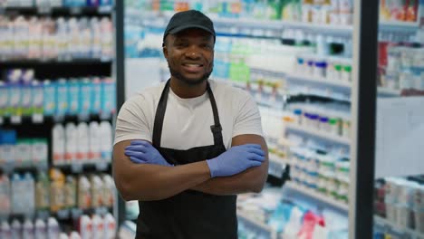 Portrait-of-a-happy-man-with-Black-skin-in-a-white-T-shirt-and-a-black-apron-who-folded-his-arms-on-his-chest-near-display-cases-with-dairy-products-in-a-large-grocery-supermarket