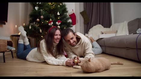 Happy-couple,-a-guy-and-a-brunette-girl-lie-on-the-floor-and-play-with-their-cream-colored-cat-with-the-help-of-a-New-Year's-toy-near-a-decorated-New-Year's-tree-in-their-cozy-home-in-winter