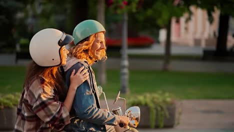 Side-view:-a-guy-with-long-curly-hair-in-a-green-moped-helmet-and-a-denim-jacket-rides-with-his-brunette-girlfriend-in-a-plaid-shirt-on-a-moped-along-the-street-at-a-golden-sunset-in-a-summer-city