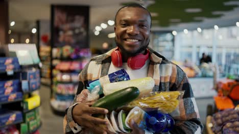 Portrait-of-a-happy-man-with-Black-skin-in-a-checkered-shirt-and-red-wireless-headphones-who-holds-a-large-amount-of-products-on-his-chest-while-holding-it-with-both-hands-in-a-modern-supermarket-near-the-display-cases