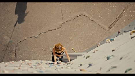 Shot-from-above,-a-blonde-girl-in-sunglasses,-a-black-sports-summer-uniform-and-in-special-equipment-for-rock-climbing-and-belaying-climbs-up-a-white-climbing-wall-with-multi-colored-ledges-and-trains-her-rock-climbing-skills-in-the-summer