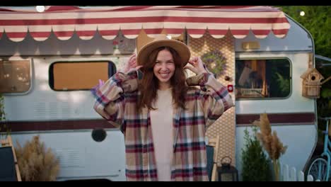 Portrait-of-a-happy-brunette-girl-in-a-brown-hat-in-a-pink-plaid-shirt-who-adjusts-her-clothes-posing-and-smiling-Near-her-trailer-outside-the-city-during-her-picnic-at-the-camp-in-the-summer