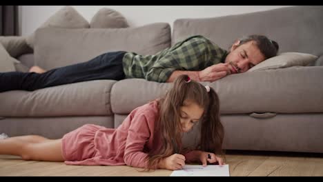 A-little-brunette-girl-in-a-pink-dress-draws-on-a-white-sheet-of-paper-with-a-pen-while-her-single-father,-a-brunette-man-in-a-green-checkered-shirt,-sleeps-on-a-gray-sofa