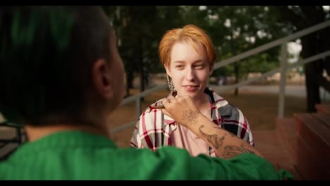 Por-Encima-Del-Hombro:-Una-Chica-Con-Cabello-Corto-Y-Verde-Con-Una-Camisa-Verde-Maquillando-A-Su-Novia-Con-Cabello-Corto-Y-Naranja-Con-Una-Camisa-Rosa-A-Cuadros-En-Bancos-Marrones-En-El-Parque