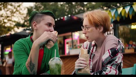 Una-Chica-Rubia-Con-Un-Corte-De-Pelo-Corto-Y-Brillo-En-Su-Cara-Junto-Con-Su-Novia-Con-Cabello-Corto-Verde-Y-Una-Camisa-Verde-Están-Sentadas-En-Una-Mesa-En-Un-Café-De-La-Calle-Y-Comen-Perritos-Calientes-Durante-Su-Cita-En-El-Parque.