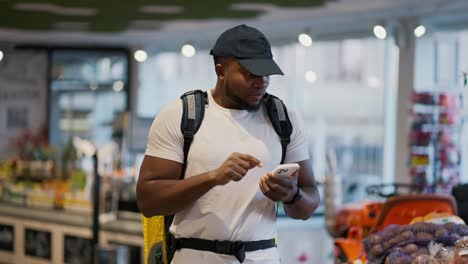 Un-Repartidor-De-Piel-Negra-Con-Camiseta-Blanca-Selecciona-Y-Busca-Los-Productos-Necesarios-Para-La-Entrega-De-Comida-Y-Sostiene-Una-Gran-Bolsa-Amarilla-Sobre-Sus-Hombros-En-Un-Supermercado-Moderno.