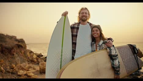 Portrait-of-a-happy-couple,-a-blond-guy-with-a-beard-in-a-checkered-shirt-stands-and-hugs-his-blonde-girlfriend-in-a-green-hat.-A-guy-and-a-girl-hold-surfboards-and-stand-on-a-rocky-seashore-in-the-morning