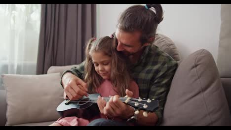 Single-father-brunette-man-with-a-green-checkered-shirt-together-with-his-little-daughter-in-a-pink-dress-playing-the-ukulele-and-tuning-a-musical-instrument-while-sitting-on-a-gray-sofa-in-a-modern-apartment