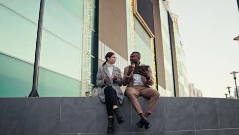 Happy-man-Businessman-with-Black-skin-in-a-brown-suit-sits-with-his-colleague-a-brunette-girl-in-a-gray-jacket-and-they-plan-their-decisions-using-a-black-tablet-while-sitting-on-a-large-gray-building-in-the-city