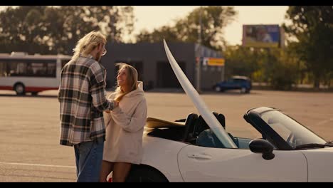 A-blond-guy-with-glasses-in-a-plaid-shirt-stands-near-his-blonde-girlfriend-in-a-white-sweatshirt-and-kisses-her,-holding-hands-near-a-white-convertible-with-a-surfboard