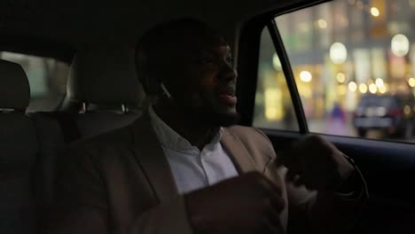 A-cheerful-and-happy-man-with-Black-skin-and-a-beard-in-wireless-headphones-listens-to-music-and-dances-while-driving-in-the-passenger-seat-in-the-modern-interior-of-a-modern-car