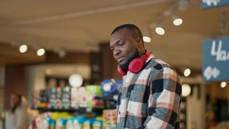 A-man-with-Black-skin-in-a-plaid-shirt-and-red-wireless-headphones-walks-through-a-modern-grocery-store-and-examines-the-goods-on-the-counter