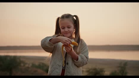 Portrait-of-a-little-blonde-girl-with-blue-eyes-in-a-white-jacket-who-peels-a-banana-and-eats-it-during-her-picnic-outside-the-city-in-the-evening-in-summer