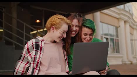 Happy-and-very-cheerful-girls-with-short-haircuts-of-bright-colors-in-checkered-shirts-and-their-friend,-a-brunette-girl,-are-looking-at-something-in-the-screen-of-a-gray-monitor-and-hugging-each-other