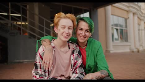 Portrait-of-two-happy-lesbian-girls-after-a-building-on-the-street-who-are-hugging-during-their-date.-A-blonde-girl-with-a-short-haircut-in-a-pink-checkered-shirt-and-a-girl-with-green-hair-in-a-green-shirt-are-hugging-during-their-date