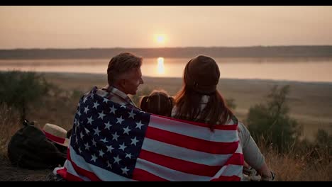 A-happy-family-wrapped-in-the-flag-of-the-United-States-of-America-sits-on-a-mat-and-watches-a-beautiful-evening-sunset-in-the-summer-during-their-picnic-outside-the-city