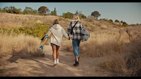 Rear-view-of-a-blond-guy-in-a-plaid-shirt-in-jeans-holding-hands-with-his-blonde-girlfriend-in-a-white-sweatshirt.-They-are-carrying-surfboards-and-walking-along-a-path-with-dried-grass-and-small-trees