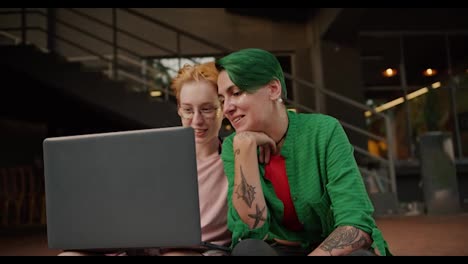 Two-lesbian-girls,-during-their-date,-look-at-the-screen-of-a-laptop-server-and-talk-about-something-near-a-large-building-in-the-summer.-A-blonde-girl-with-a-short-haircut-and-a-girl-with-green-hair-with-a-short-haircut-in-a-green-shirt-sit-and-look-at-the-laptop-screen-during-their-date-and-make-t