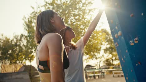 A-blonde-girl-in-black-glasses-and-a-white-T-shirt-tells-her-friend-Rock-girl-with-a-bob-hairstyle-in-a-black-top-how-they-will-climb-a-route-at-a-climbing-wall-on-a-sunny-day-in-the-summer