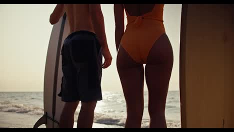 Zoom-in:-A-brunette-guy-in-shorts-and-a-blonde-girl-in-an-orange-swimsuit-stand-next-to-each-other-and-hold-surfboards-and-then-they-hold-hands-and-look-towards-the-sea