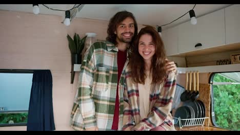 Portrait-of-a-happy-couple,-a-brunette-guy-with-stubble-in-a-green-checkered-shirt-hugs-his-brunette-girlfriend-in-a-pink-checkered-shirt-in-their-campervan-outside-the-city-in-a-camp-during-a-picnic-in-the-summer