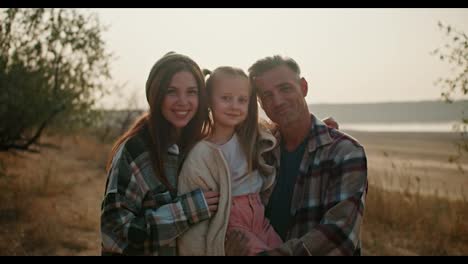 Portrait-of-a-happy-family,-a-brunette-man-with-gray-hair-in-a-checkered-shirt-poses-with-his-little-daughter-and-his-wife,-a-brunette-girl-in-a-green-checkered-shirt-against-of-nature-outside-the-city-with-dry-grass-and-small-green-trees