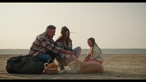 Happy-brunette-man-with-gray-hair-in-a-checkered-shirt-pets-his-dog-and-relaxes-with-his-wife-and-daughter-on-his-picnic-during-a-hike-on-a-deserted-seashore-in-summer