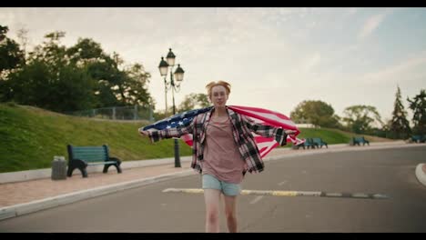 A-blonde-girl-with-short-hair-with-glasses-in-a-checkered-pink-shirt-runs-with-the-flag-of-the-United-States-of-America-behind-her-shoulders-along-the-road-in-the-park-in-the-summer