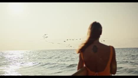 Rear-view-of-a-blonde-girl-in-an-orange-swimsuit-standing-with-a-surfboard-on-the-seashore-and-looking-at-the-sea-and-the-sky-where-a-large-flock-of-birds-are-flying-at-Sunrise-in-the-morning