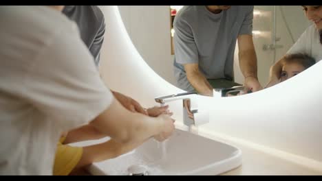 Zoom-in:-Little-girl-and-her-parents-wash-their-hands-under-running-water-in-a-modern-sink-near-the-mirror-in-the-bathroom