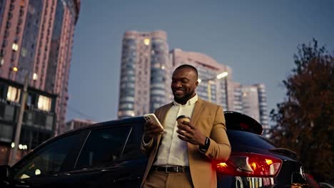 A-happy-and-joyful-Black-man-with-a-short-haircut-and-a-beard-is-using-a-white-phone-and-drinking-coffee-in-a-brown-suit-near-his-modern-black-car-in-a-city-with-large-buildings-and-a-blue-sky-in-the-evening