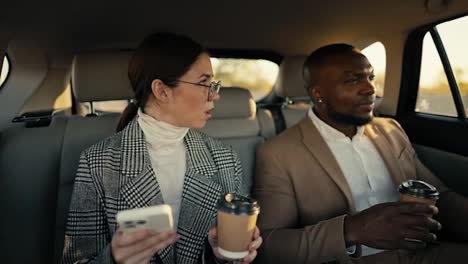 A-confident-brunette-businesswoman-in-round-glasses-with-a-cup-of-coffee-in-her-hands-and-a-smartphone-communicates-with-her-colleague-a-man-with-Black-skin-and-a-brown-jacket-during-his-business-trip-in-the-cabin-of-a-modern-car-outside-the-city