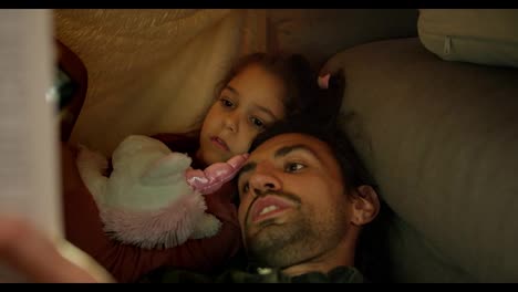 Close-up-shot-of-a-brunette-man-in-a-green-checkered-shirt-reading-a-fairy-tale-to-his-daughter-under-a-makeshift-awning-at-home-near-a-gray-sofa.-A-little-brunette-girl-lies-under-a-makeshift-awning-at-home-and-listens-to-her-father’s-stories-with-a-unicorn-toy-in-her-hands