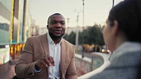 Over-the-shoulder-A-confident-man-with-Black-skin-with-a-short-haircut-and-a-beard-in-a-brown-jacket-talks-about-his-plans-and-decisions-in-the-upcoming-quarter-during-a-business-meeting-with-a-businesswoman-girl-in-the-city