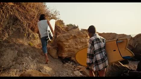 A-brunette-guy-in-a-white-T-shirt-together-with-his-girlfriend-in-a-Green-hat-and-a-plaid-shirt-walk-along-the-rocky-shore-along-the-sea-and-carry-surfboards-with-them-on-an-autumn-morning