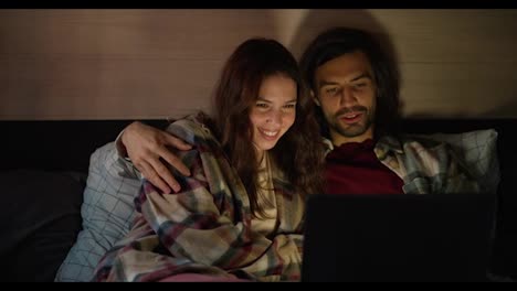 Happy-brunette-girl-in-a-plaid-shirt-lies-on-the-sofa-in-a-trailer-with-her-boyfriend-with-stubble-and-they-watch-a-movie-together-using-a-laptop-during-their-vacation-in-a-camp-outside-the-city-in-the-summer-with-dim-lights