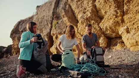 A-blonde-girl-with-curly-hair-in-a-white-T-shirt,-a-blonde-girl-with-a-bob-hairstyle-in-a-Blue-jacket-and-a-brunette-guy-in-a-gray-T-shirt-are-sorting-out-their-backpacks-and-preparing-equipment-for-rock-climbing-on-a-rocky-seashore-near-the-yellow-rocks-on-a-summer-day