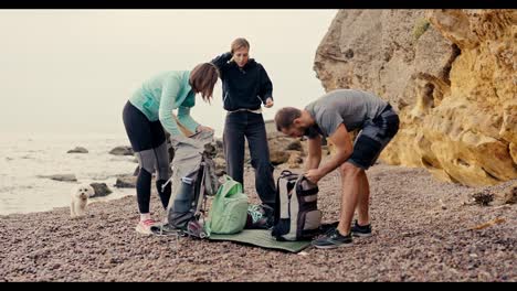 A-group-of-rock-climbers-unpack-their-backpacks-and-prepare-to-climb-the-yellow-rocks-near-the-sea-on-a-rocky-shore-in-summer.-A-blonde-girl-in-a-black-sports-uniform,-as-well-as-a-girl-with-a-bob-hairstyle-and-a-brunette-guy-are-taking-apart-their-backpacks-and-preparing-to-climb-the-rocks