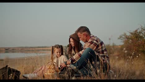 Side-view-of-a-happy-family-having-a-picnic-outside-the-city-near-dry-grass-and-lonely-small-green-trees-near-a-deserted-seashore-in-summer.-A-brunette-man-with-gray-hair-in-a-brown-checkered-shirt-pours-tea-for-his-wife-and-little-daughter-from-a-thermos-during-his-picnic-and-vacation-outside-the-c