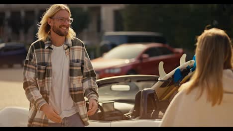 A-happy-blond-guy-with-a-beard-in-a-checkered-shirt-meets-his-blonde-girlfriend-in-a-white-sweatshirt,-who-runs-towards-him.-Happy-couple-meeting-near-a-convertible-on-a-sunny-summer-day