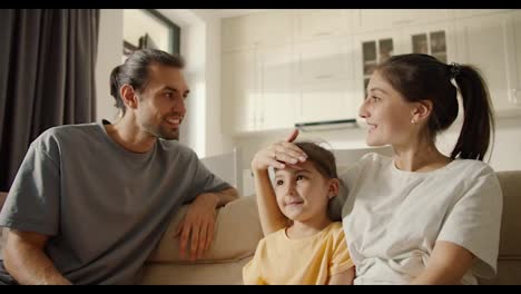 Well,-I'm-a-happy-brunette-guy-in-a-gray-T-shirt-talking-with-his-brunette-girlfriend-and-their-daughter,-a-brunette-girl-in-a-white-T-shirt,-sitting-on-a-brown-sofa-in-a-modern-studio-room