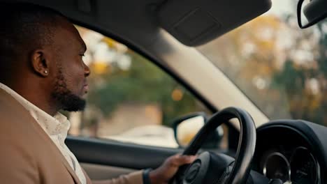 Vista-Lateral-De-Un-Hombre-De-Negocios-Seguro-De-Sí-Mismo,-De-Piel-Negra-Y-Barba-Con-Una-Chaqueta-Marrón,-Conduce-Un-Automóvil-Durante-Su-Viaje-De-Negocios-En-Una-Ciudad-Moderna-Y-Mira-Por-La-Ventana-Lateral.