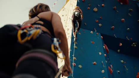 Shot-from-below,-a-blonde-girl-in-a-black-sports-summer-uniform-with-the-necessary-equipment-and-insurance-climbs-up-a-blue-climbing-wall,-and-a-girl-with-a-bob-hairstyle-in-a-black-summer-uniform-insures-her-and-holds-the-necessary-rope-to-support-her-friend