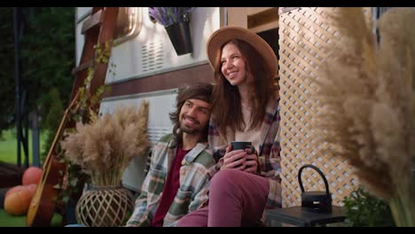 Happy-brunette-girl-in-a-light-brown-black-mug-with-a-hot-drink-in-her-hands-and-communicates-with-her-brunette-boyfriend-in-a-green-checkered-shirt-sitting-near-a-decorated-trailer-in-a-camp-during-a-picnic-outside-the-city-in-the-summer