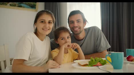 Portrait-of-a-happy-and-joyful-family,-a-brunette-man-in-a-gray-T-shirt,-his-wife,-a-girl-in-a-white-T-shirt,-and-their-little-daughter,-a-brunette-girl-in-a-yellow-dress,-sitting-at-the-family-white-table-and-breakfast-lying-on-plates-in-the-kitchen-in-the-morning