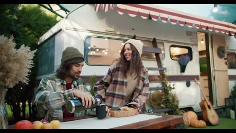 A-happy-brunette-girl-comes-out-of-the-trailer-and-brings-out-delicious-pastries-to-her-boyfriend-while-He-pours-tea-and-chats-with-his-girlfriend-while-sitting-at-a-table-near-the-trailer-outside-the-city-during-a-picnic-at-the-camp-in-the-summer